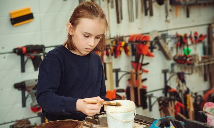 Stock-foto fra Colourbox. En elev står med træolie og pensel i et håndværkslokale, hvor værktøj hænger i baggrunden. 