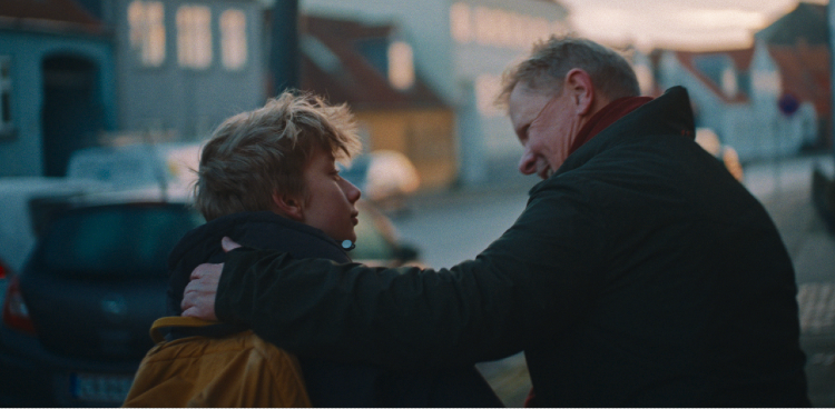 Fotoet viser en dreng sammen med en mand bagfra. Drengen har skoletaske på og manden har lagt armen på ryggen af drengen og sender ham et smil. Foto: Lær for Livet