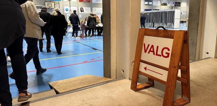 Billedet viser borgere i kø til stemmeafgivelse på et lokalt valgsted i en sportshal. I forgrunden af billedet ses et skilt med en pil og teksten "valg". Foto taget af Skanderborg Kommune