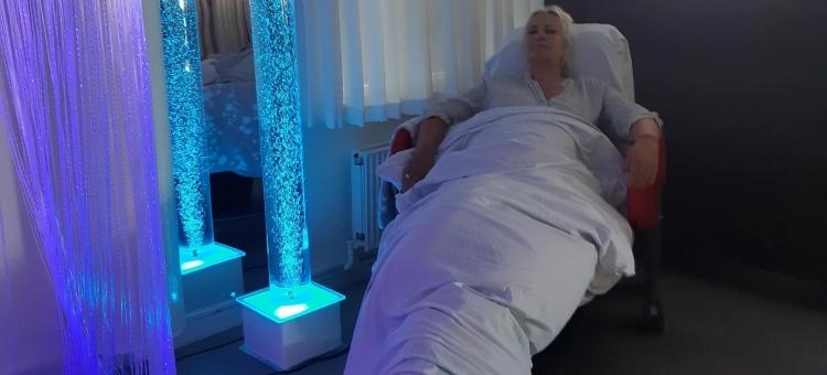 Billede af dame i liggestol med dyne over sig med en boblelampe foran sig, der lyser blåt. 
