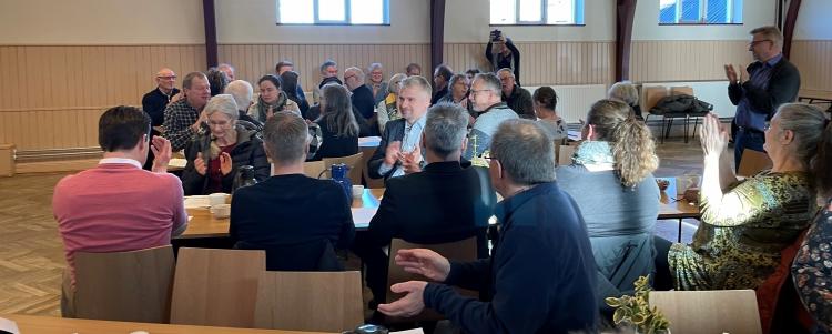 Foto af en række mennesker samlet i forsamlingshus. Mange markerer med hånden i vejret. 