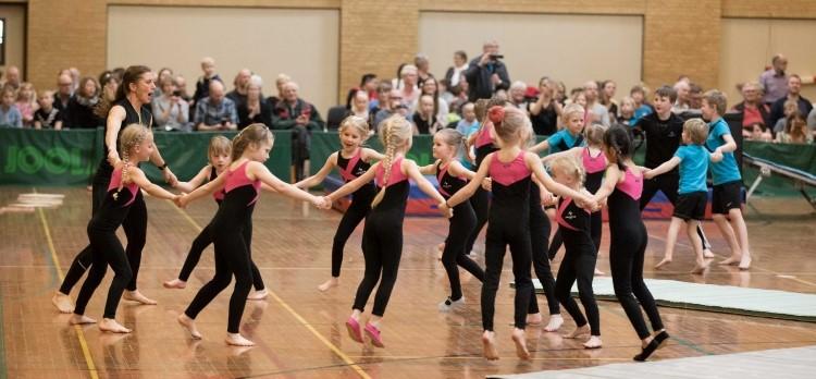 Børn i gymnastikdragter og hinanden i hånden hopper rundt i rundkreds i en hal med tilskuere.