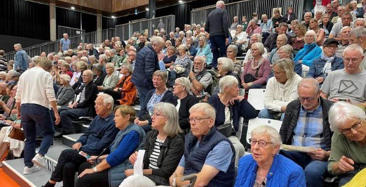 Mange seniorer sidder på tribunen i hallen på Fælleden og er klar til foredrag. 