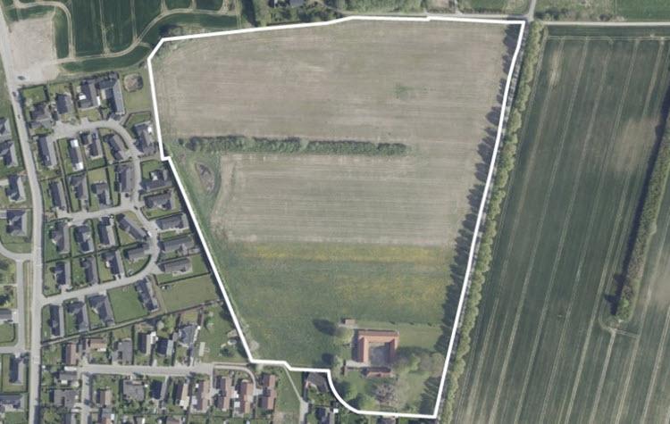 Luftfoto af området, der er omkranset af en hvid markering. I nærheden ses parcelhuse og en mark. 