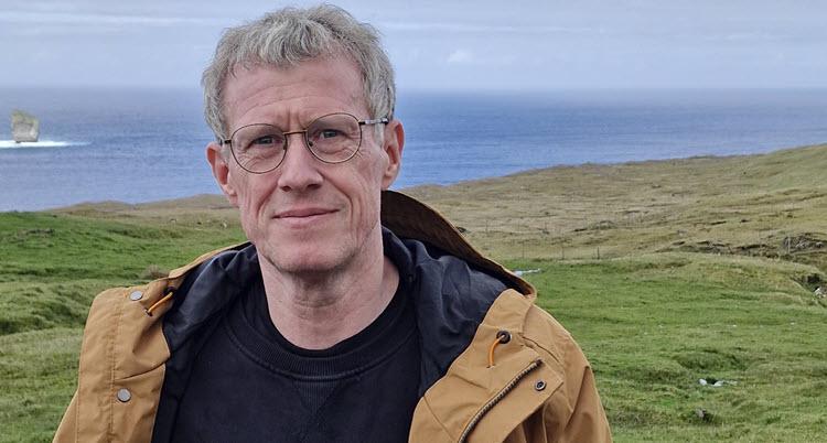En mand i en brun jakke står i et naturområde med grønne marker og havet i baggrunden. 