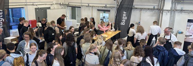 Et stort lokale fyldt med 7. klasses-elever, der besøger forskellige fagboder til Mini Skills i Skanderborg. I forgrunden ses elever i kø ved en stand med skiltet “Bager”, hvor fagpersonale hjælper dem i gang med opgaver.