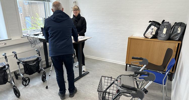 En mand med stok står over for en kvindelig medarbejder ved et hæve-sænkebord i et lyst lokale. Rundt om står to rollatorer, en rollatorkurv og hjælpemiddeltasker. 