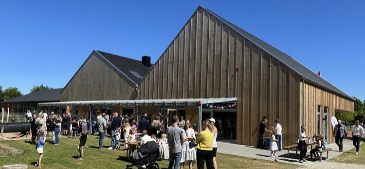 Den nybyggede institution Stjær Børnehus med høje saddeltage og facade i lodrette trælameller. Foran bygningen samles børn og voksne i solskinnet på en græsplæne og legeplads..