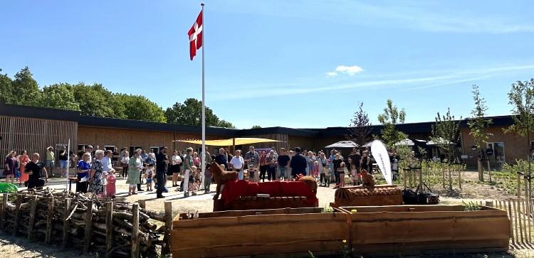 Gæster står samlet under Dannebrog foran det nye børnehus med træfacade og nyplantede træer.