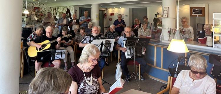 Et kor af ældre borgere synger, mens nogle spiller guitar og harmonika i caféens fællesrum.