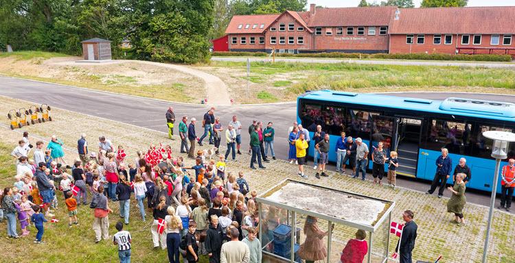 Dronefoto af en masse børn og voksne samlet på en parkeringsplads med et busskur og en blå bus. I baggrunden ses et nybygget toilethus, Låsbyvej og Veng Skole.