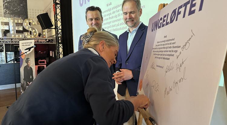 En kvinde skriver under på en planche med overskriften "Ungeløftet", mens borgmesteren og finansministeren ser smilende til.