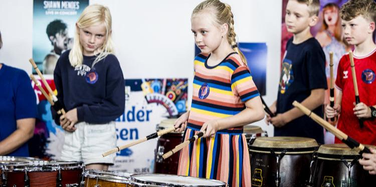 En række børn står ved forskellige trommer på en scene med plakater bagved. 