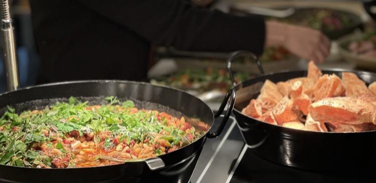 Frokostanretning med stor gryde med bønner og grønt i forgrunden og i baggrunden er en hånd, der tager salat op på en tallerken.