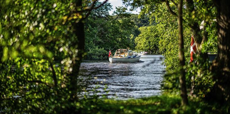 Lille båd med Dannebrogsflag sejler på Gudenåen. Billedet er taget fra land gennem træer med grønne blade. 