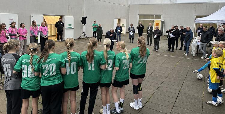 Børn i idrætstøj og med bolde står mellem to halbygninger, mens en mand taler i mikrofon og mange mennesker lytter.