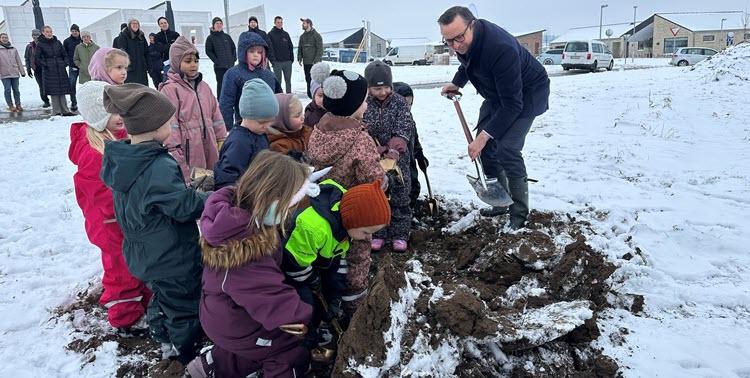 Borgmester Frands Fischer sammen med en række børn i flyverdragter graver i jord med sne og tilskuere i baggrunden. 