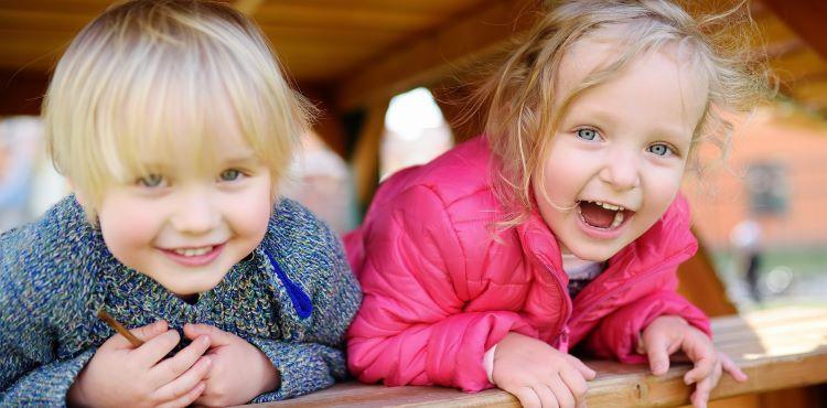 En dreng og pige i børnehavealderen gemmer sig under underdørs bord-bænkesæt og smiler til fotografen.
