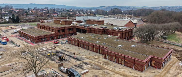 Dronefoto af byggepladsen med en 1-etages bygning og en bygning på flere etager i forgrunden. I baggrunden ses Ry Hallerne og Søhøjlandets bakkede og skovklædte landskab.
