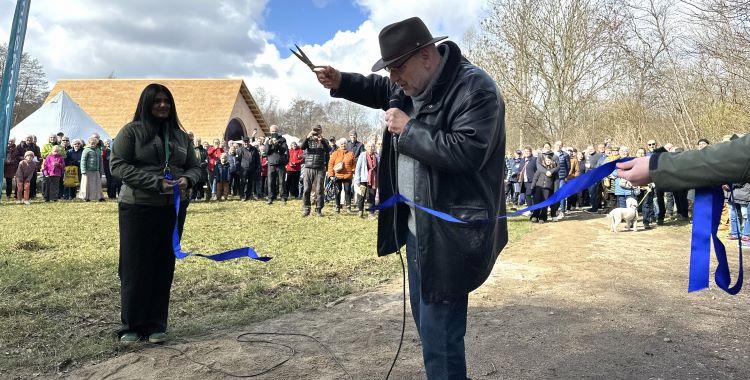 En mand med en guldsaks klipper en blå snor, som går henover en sti, foran en stor menneskemængde. I baggrunden ses den store træbygning Skimming Bådhus. 