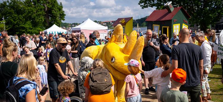 Foto af et gult næsehorn (kostume), som en masse børn og voksne er i gang med at hilse på. I baggrunden er mange mennesker og farverige træhuse og boder.