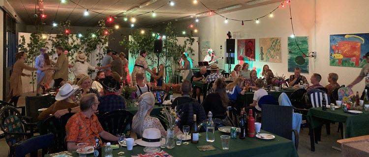 Mennesker samlet til fest i et lokale med kulørte lamper og grønne grene. Nogle sidder ved bordene, mens andre danser i nærheden af orkestret.