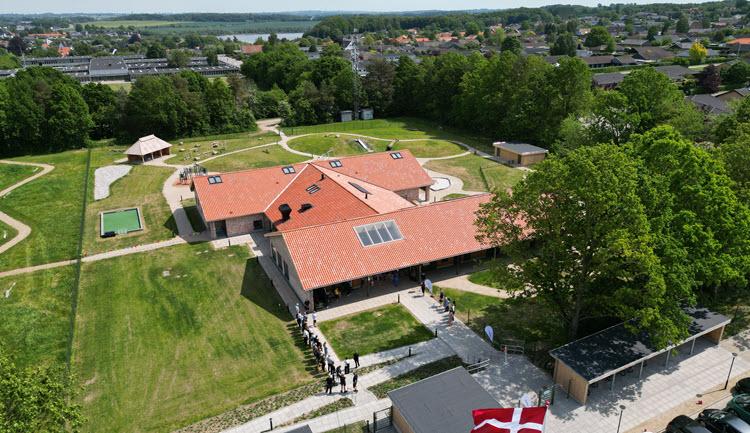 H-formet bygning med rødt tag og ovenlysvinduer med legeplads og grønne områder omkring. I forgrunden er Dannebrog hejst, og der står mennesker og lytter. I baggrunden er der træer og Stilling Skole.