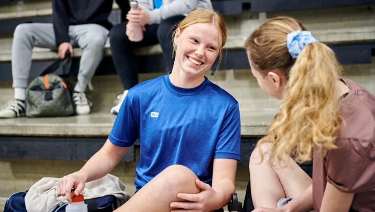 En ung kvinde i shorts og blå DGI-sportstrøje snakke med en anden kvinde i en sportshal. I baggrunden ser man to andre med sportstøj.