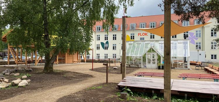 Foto af FLOK, en stor hvid bygning med store farvede sommerfugle og blomster på facaden. I forgrunden ses et lille sceneområde, et overdækket mødested og et orangeri.