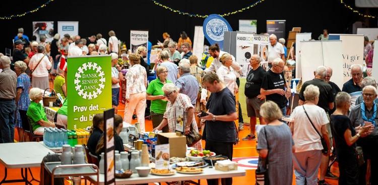Billede fra 2024 som viser en fyldt hal med boder og seniorer og roll-ups. Fotograf: Per Bille