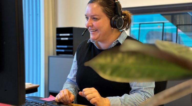 En smilende kvinde med skjorte, vest og headset sidder ved en computer på et kontor. 