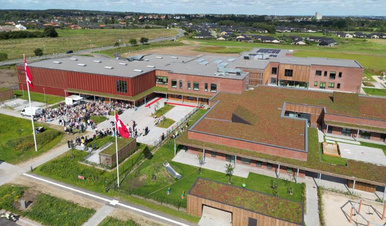 Dronebillede fra indvielsen af Sommerbæk Børnehus og Skole. Flaget er hejst, der er røde løbere og en menneskemængde. Høring kan ses i horisonten.