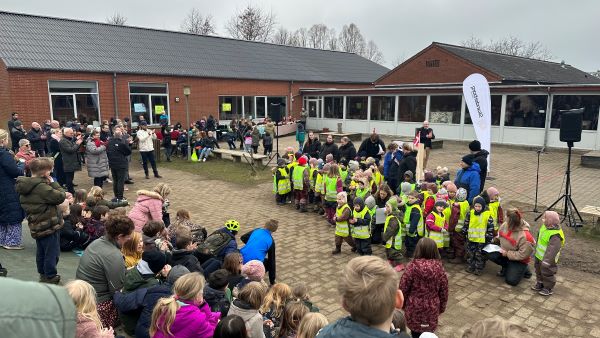 Børn i gule veste synger og publikum klapper ved en rød skolebygning.