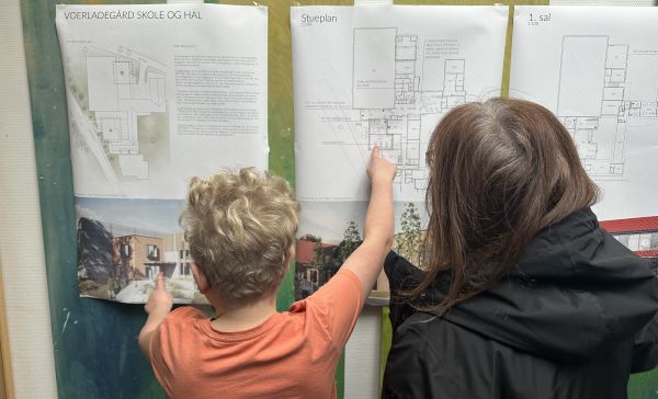 Et barn i orange tshirt peger på byggetegninger klistret op på en væg, mens en voksen kigger på.